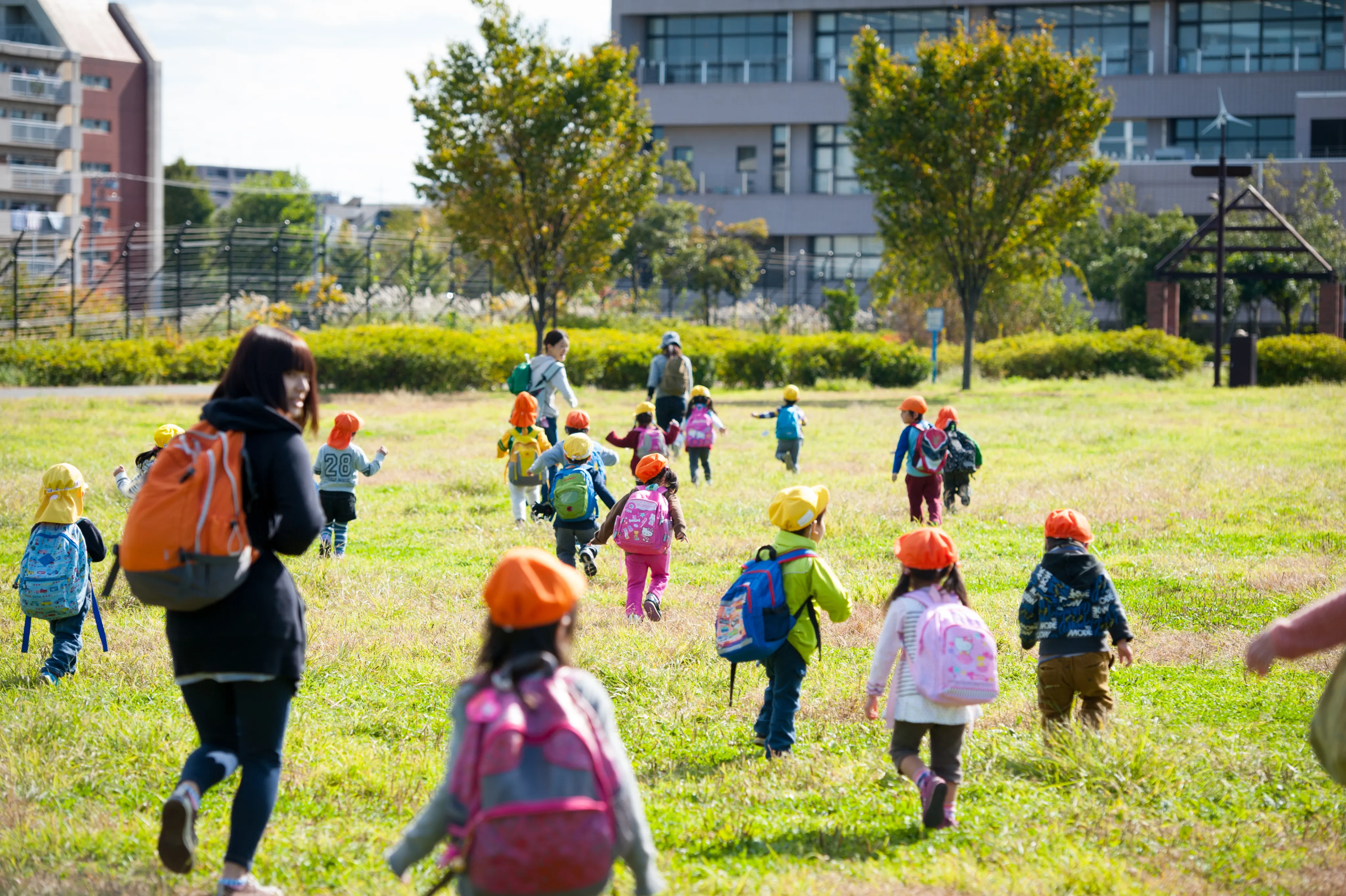 色とりどりの帽子をかぶった園児たちが、保育士と一緒に広い芝生の上を元気に歩いている様子