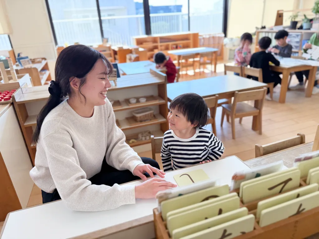 保育士と園児が笑顔で見つめ合いながら、数字のカードを使って活動している様子