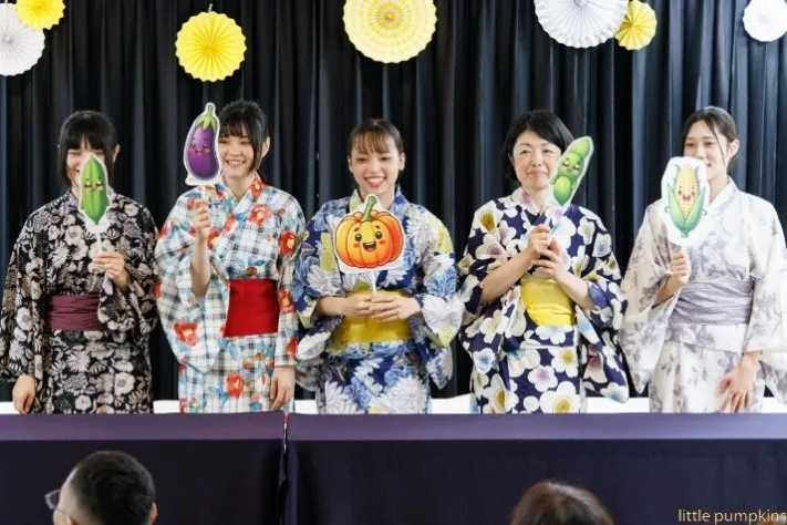 浴衣姿の保育士たちが野菜のイラストを持って笑顔で並んでいる様子