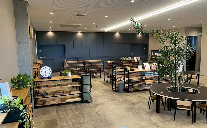 観葉植物が中央に置かれた円形テーブルや木製家具が並ぶ、落ち着いた雰囲気の保育施設の室内